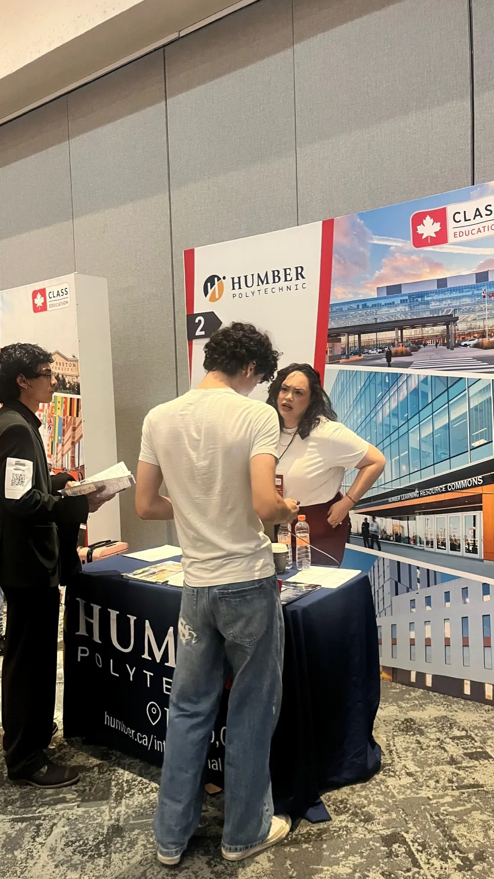 Un estudiante conversa con la representante de Humber Polytechnic en su stand mientras otro joven hojea un folleto.