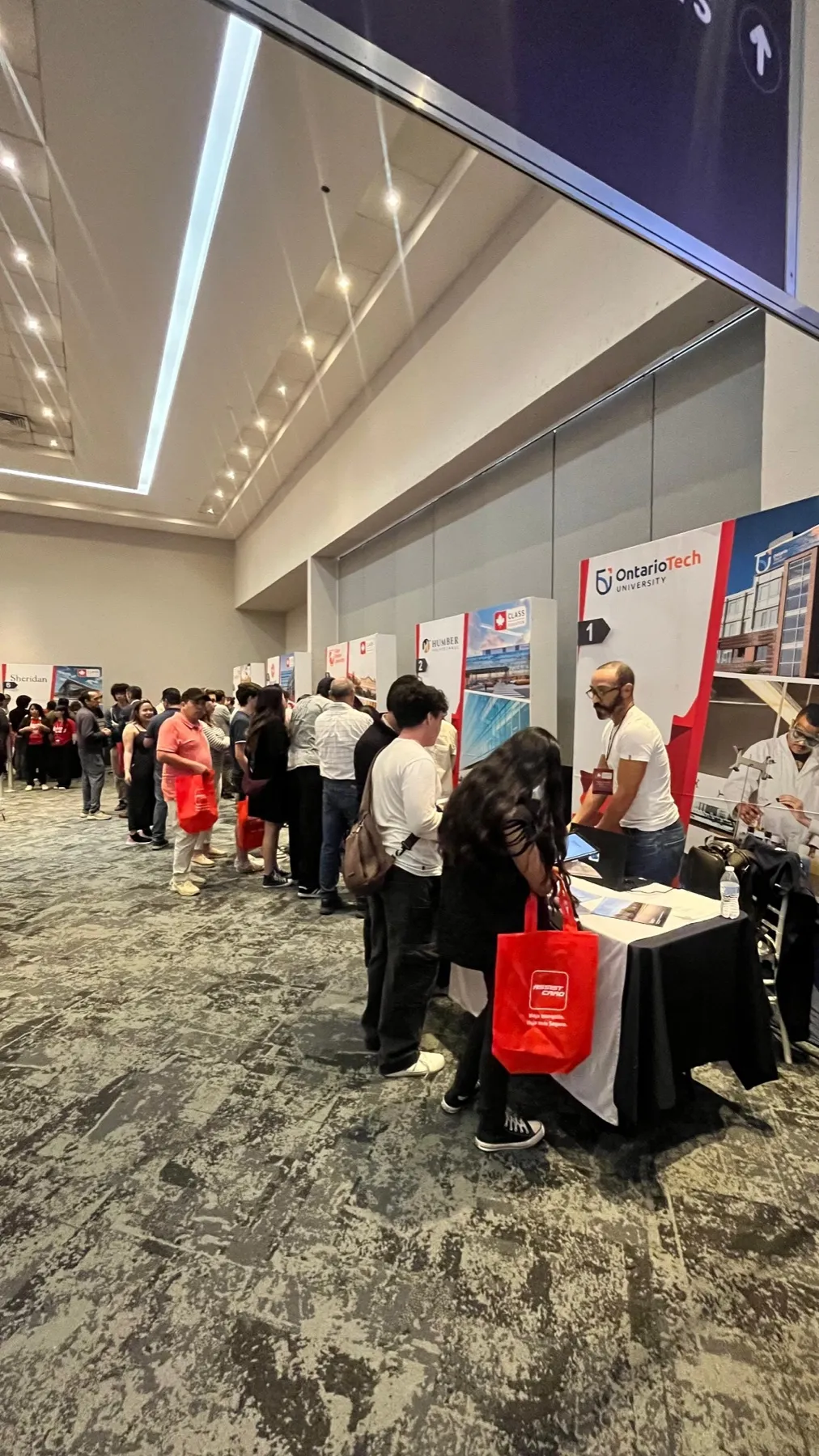 Pasillo de stands lleno de visitantes, varios con bolsas rojas de la expo; al frente, el stand de Ontario Tech University.