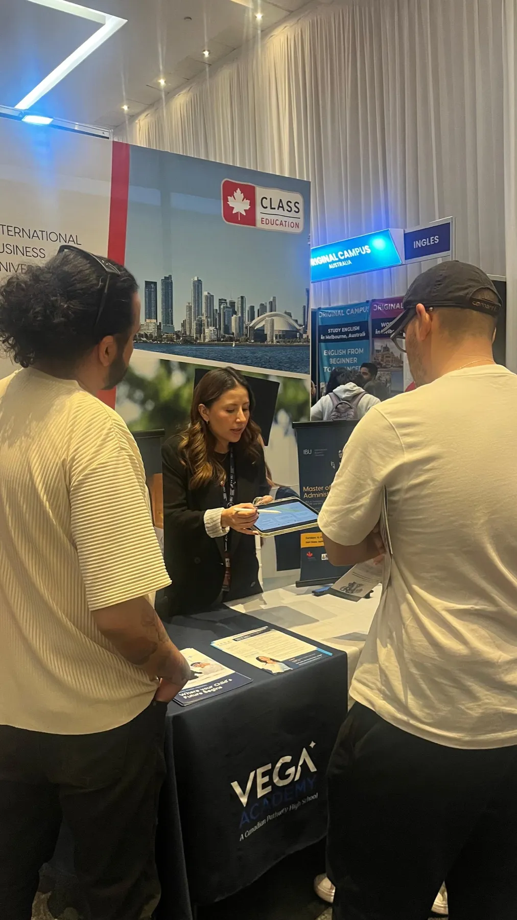 Una asesora muestra información en una tableta a dos jóvenes en el stand de Vega Academy e International Business University.